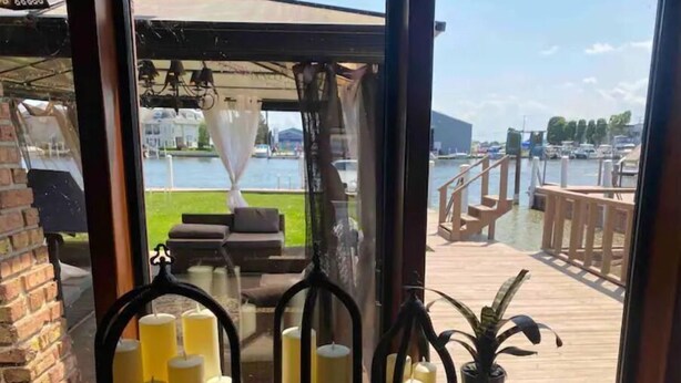 A view from a room with a table displaying candles and a potted plant, overlooking a wooden deck and a body of water.