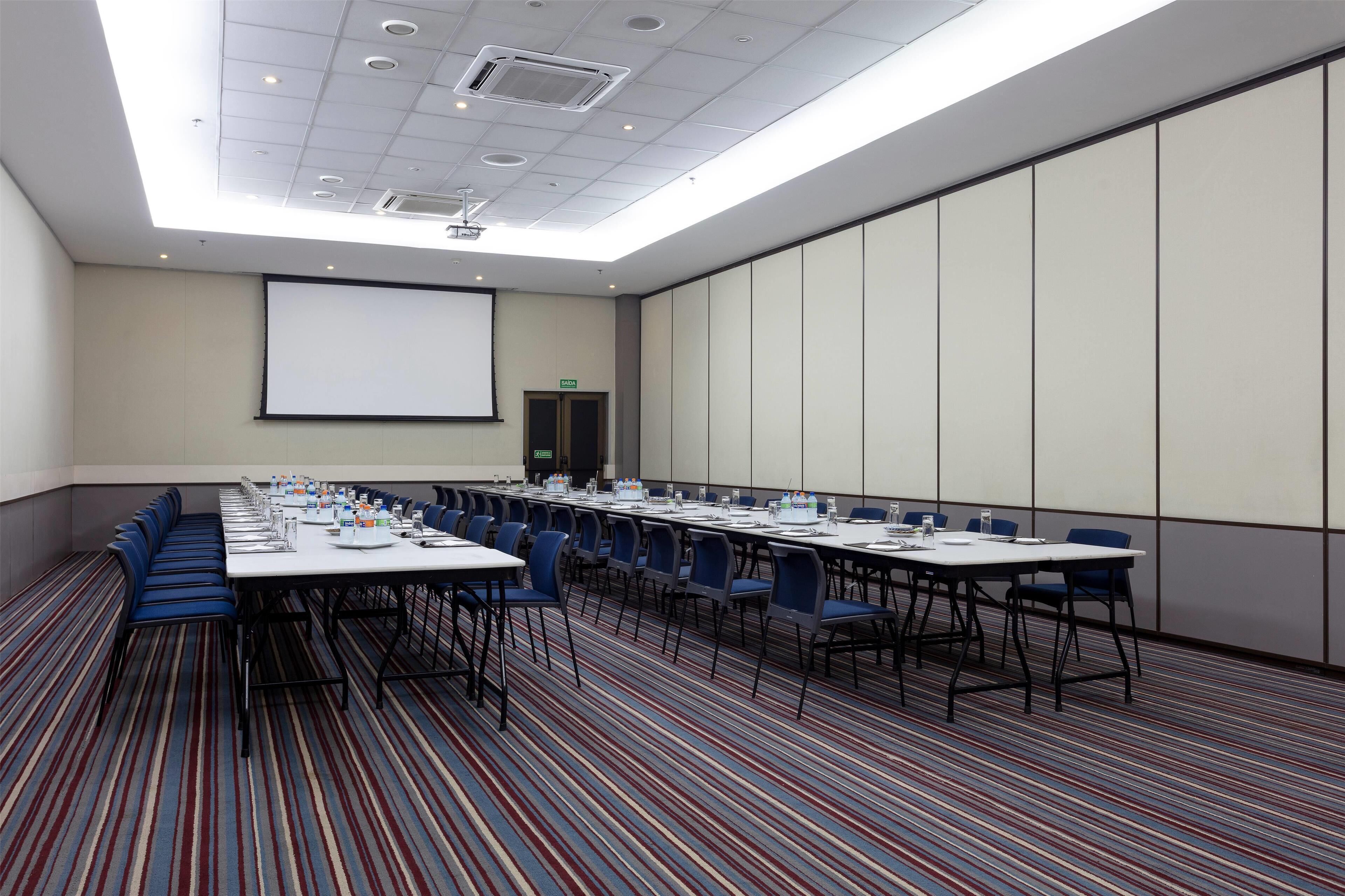 Un salón de conferencias con una larga mesa, sillas azules y una pantalla para proyector.