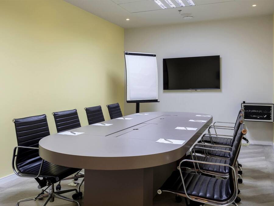 A conference room with a large table, six chairs, a projector screen, and a TV on the wall.