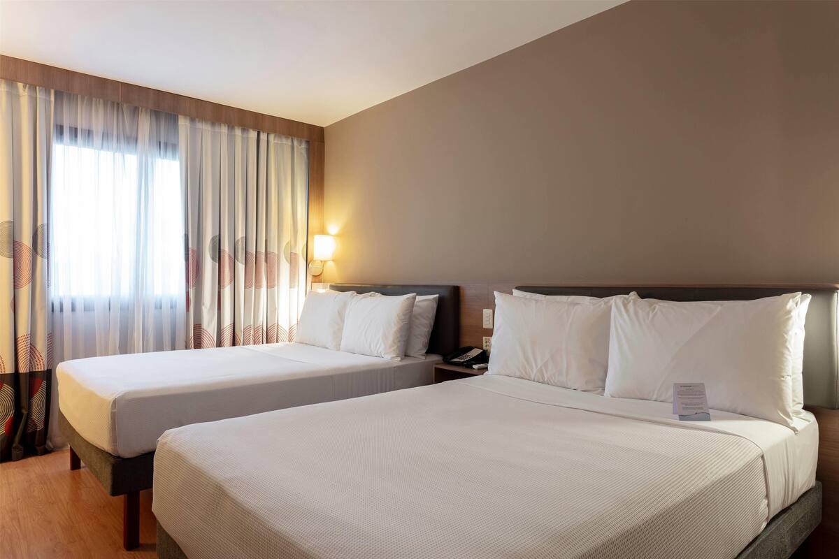Une chambre d’hôtel avec deux lits, une table de chevet, une lampe et une fenêtre avec des rideaux.