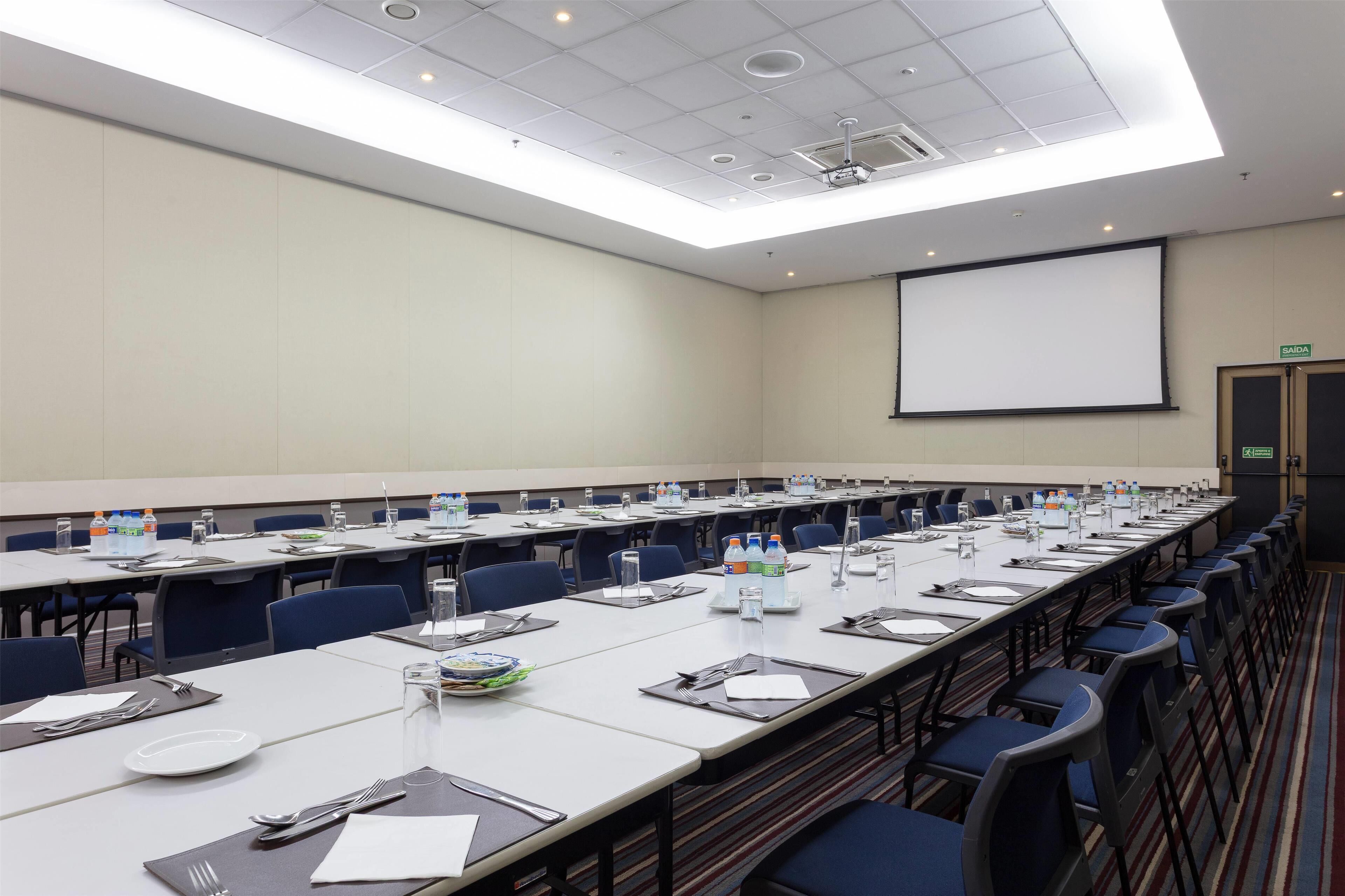 A conference room with long tables, chairs, water bottles, and a projector screen.