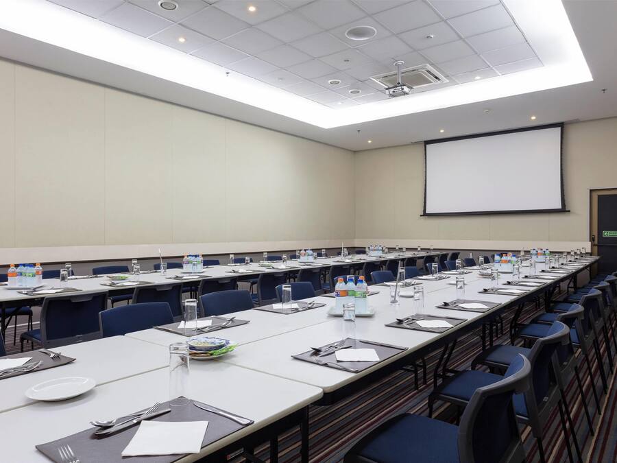 A conference room with long tables, chairs, water bottles, and a projector screen.