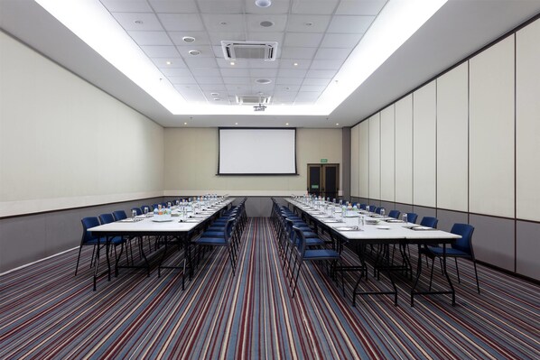 Un salón de conferencias con una larga mesa, sillas azules, una pantalla de proyector y piso alfombrado.