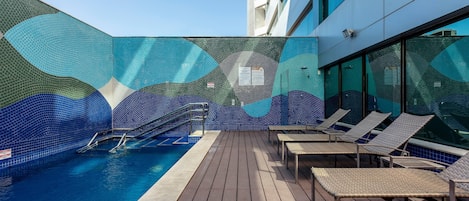 A pool area with a tiled wall, lounge chairs, and a wooden deck.