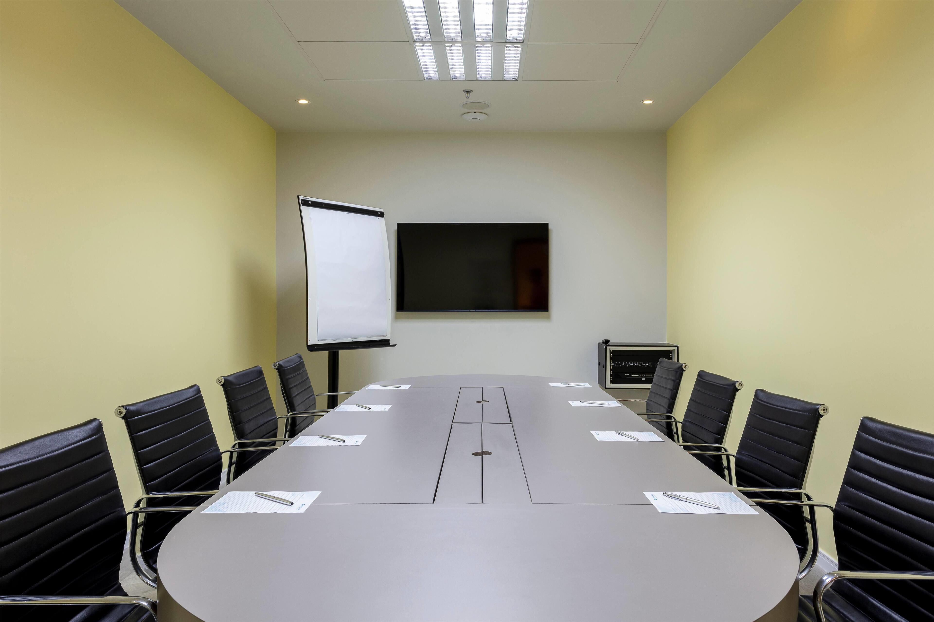 Une salle de conférence avec une longue table, des chaises noires, un tableau blanc et un écran de projection.