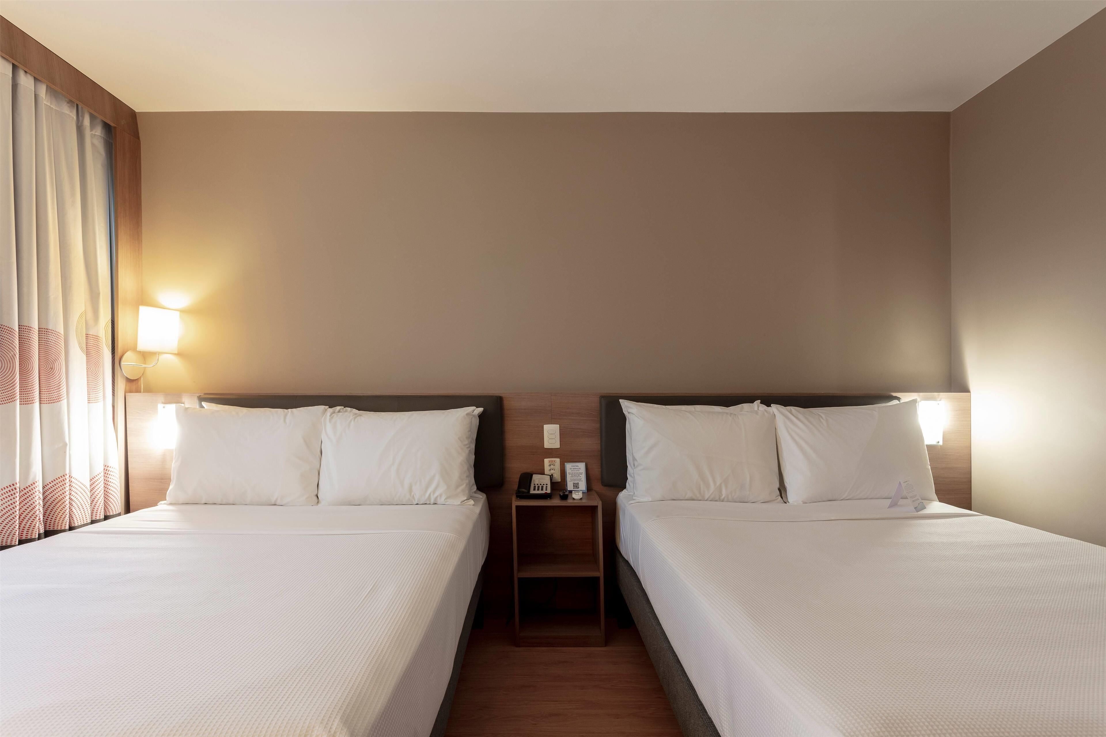 A hotel room with two single beds, a nightstand with a lamp, and a window with curtains.
