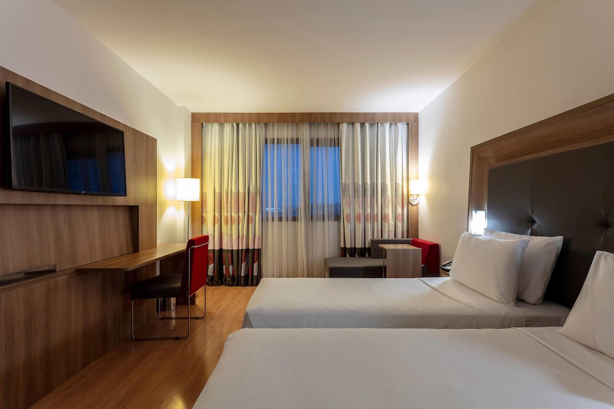 Une chambre d’hôtel avec deux lits, un bureau, une chaise, une télévision et une fenêtre avec des rideaux.