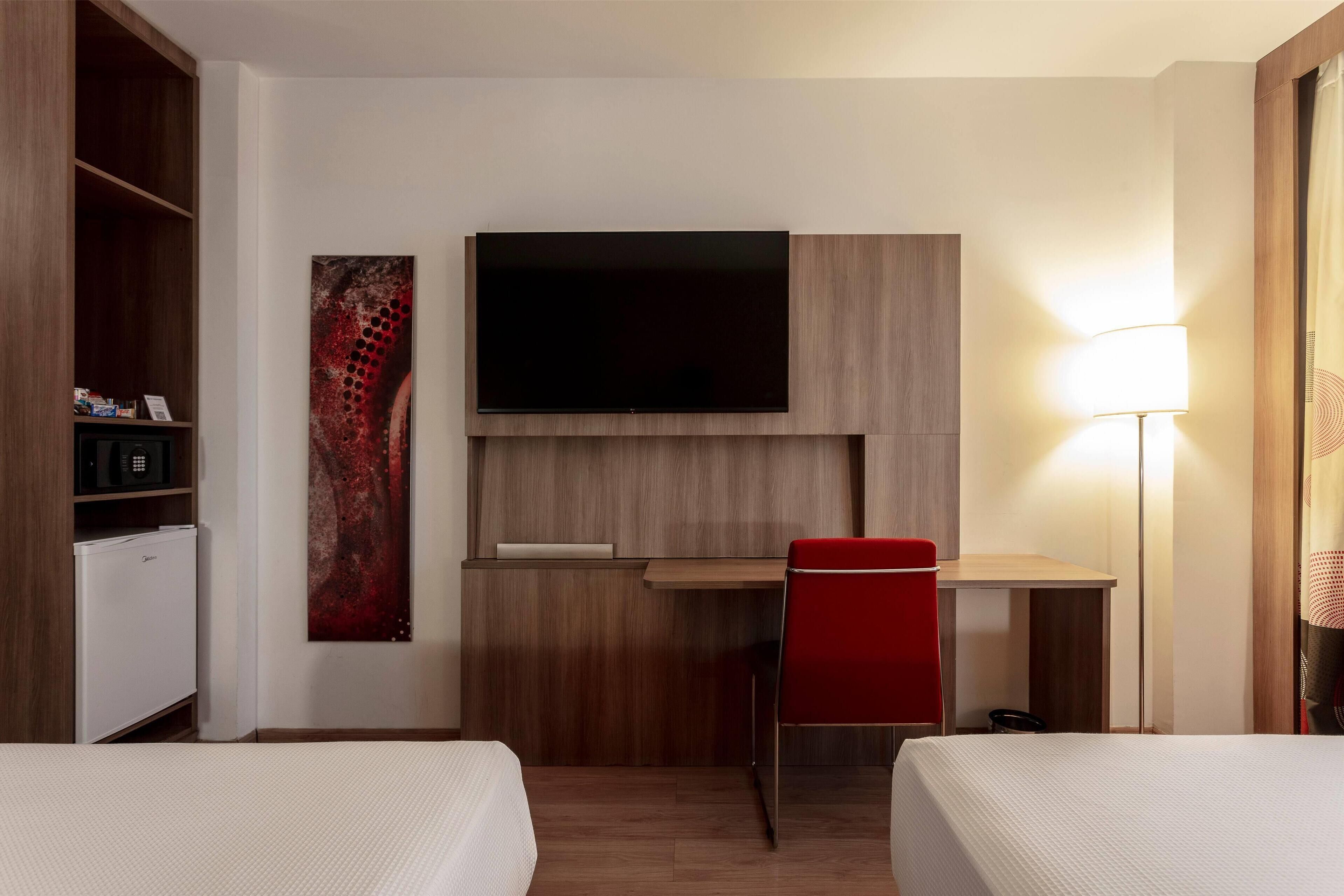 A hotel room with a bed, a desk, a TV, a red chair, and a lamp.