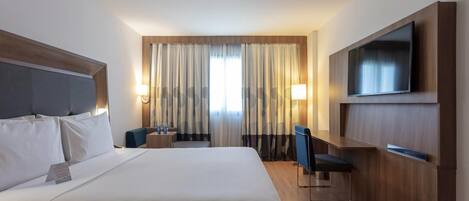 A hotel room with a large bed, a desk, a chair, a TV mounted on the wall, and curtains on the windows.