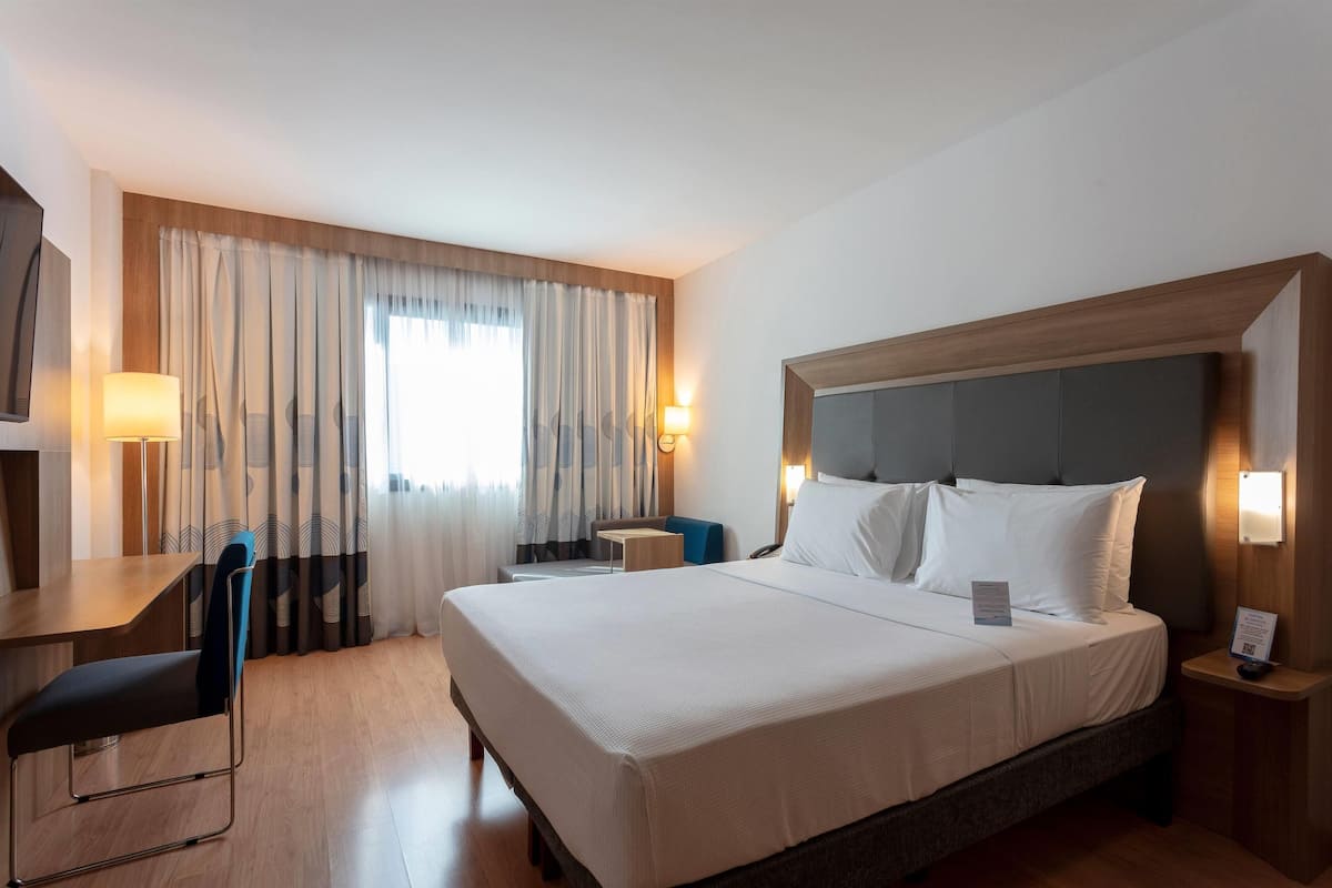 Une chambre d’hôtel avec un grand lit, un bureau et une chaise. Il y a des rideaux à la fenêtre et une lampe sur le bureau.