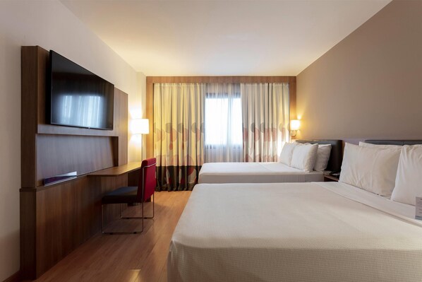 A hotel room with two beds, a TV mounted on the wall, a desk with a chair, and a window with curtains.