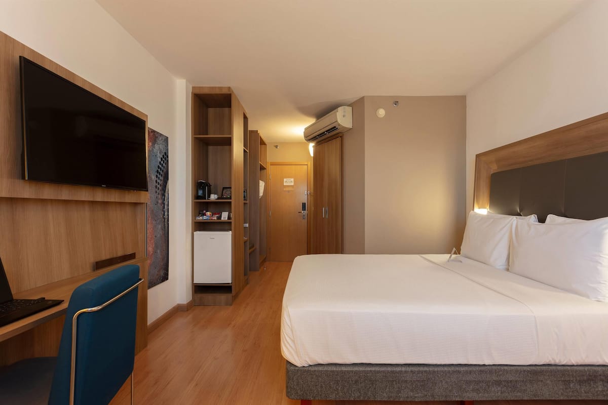 Une chambre d’hôtel avec un grand lit, un bureau avec un ordinateur portable, un téléviseur fixé au mur et une kitchenette.