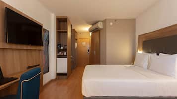 A hotel room with a large bed, a desk with a laptop, a TV mounted on the wall, and a kitchenette.
