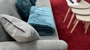 Un sofá gris con cojines azul verdosos y grises, una manta azul y una mesita redonda con un florero y un control remoto.