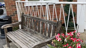 Un banco de madera desgastada con estructura metálica, ubicado frente a un muro de ladrillo con una ventana y una maceta con flores rojas y blancas.