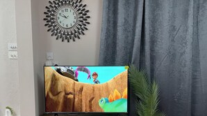 Un televisor sobre un soporte de madera, un reloj de pared y una cortina.