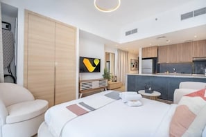 A modern hotel room with a large bed, a sitting area, a kitchenette, and a TV mounted on the wall.