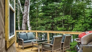 Una terraza con mobiliario de mimbre y una parrilla, rodeada de un bosque.