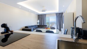 A modern hotel room with a kitchenette, a dining area with a table and chairs, a sofa, and a view of the outdoors. - Sunset Harbour Costa Adeje Beautiful 2 bedrooms apartment with amaizing sea view (Tenerife)