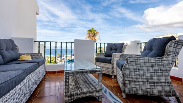 A balcony with wicker furniture, a glass table, and a view of the beach and palm tree. - Sunset Harbour Costa Adeje Beautiful 2 bedrooms apartment with amaizing sea view (Tenerife)