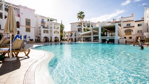 A swimming pool with clear blue water, surrounded by white buildings with balconies and palm trees. - Sunset Harbour Costa Adeje Beautiful 2 bedrooms apartment with amaizing sea view (Tenerife)