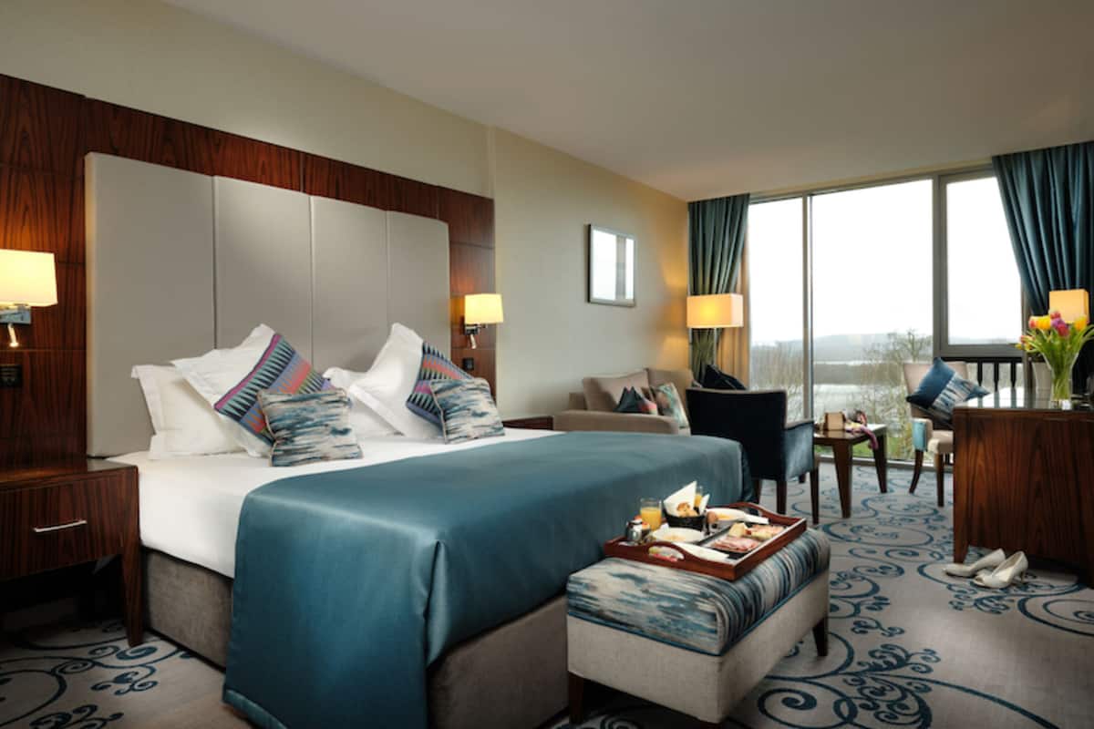A hotel room with a large bed, a sitting area with a chair and table, and a view of the outdoors through the window.
