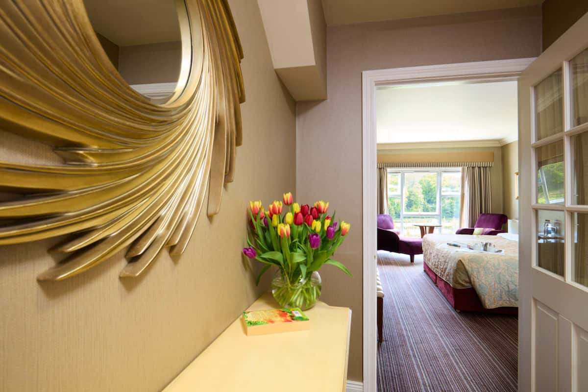 A hotel room with a bed, a chair, a desk with a vase of flowers, and a mirror on the wall.