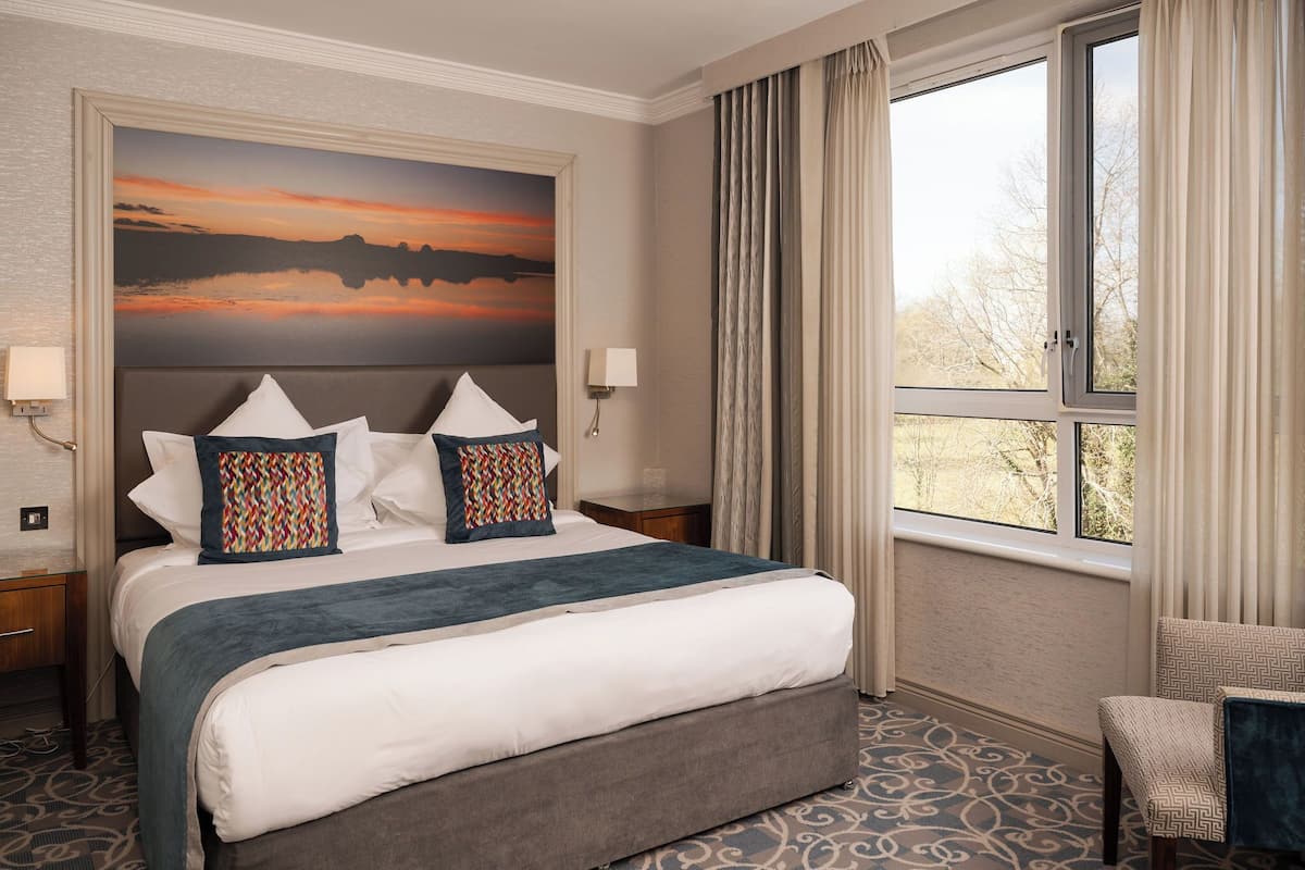 A hotel room with a large bed, a painting of a sunset, a window with curtains, and a chair.
