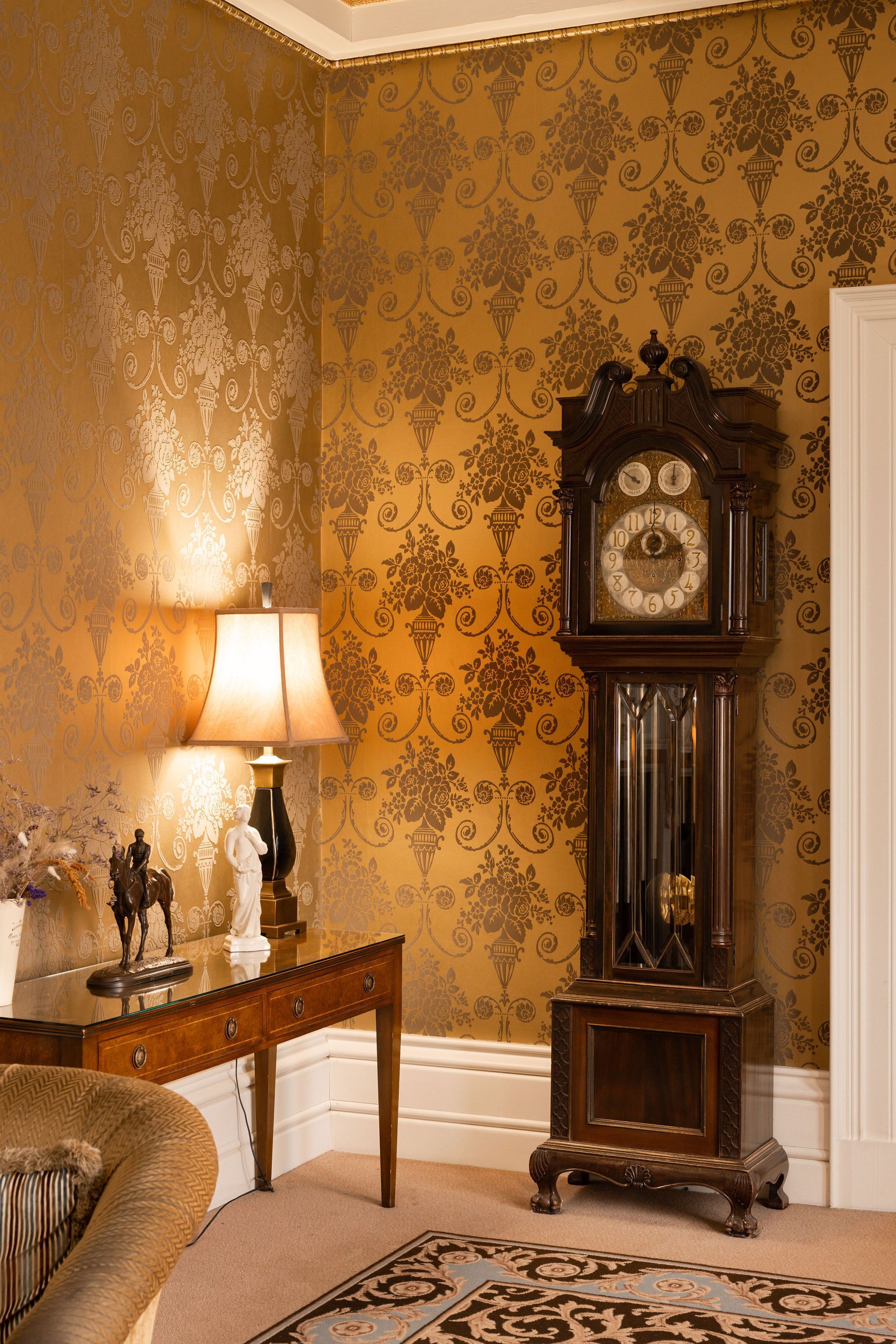 Une pièce avec un papier peint à motifs, une table en bois avec une lampe, une horloge et un canapé.