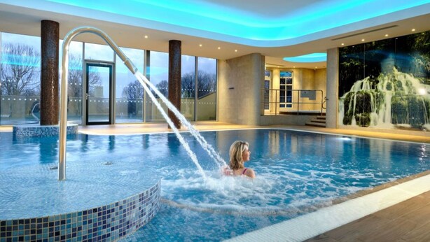 A swimming pool with a water feature, a glass door entrance, and a view of trees outside. - Manor House Country Hotel (Enniskillen)