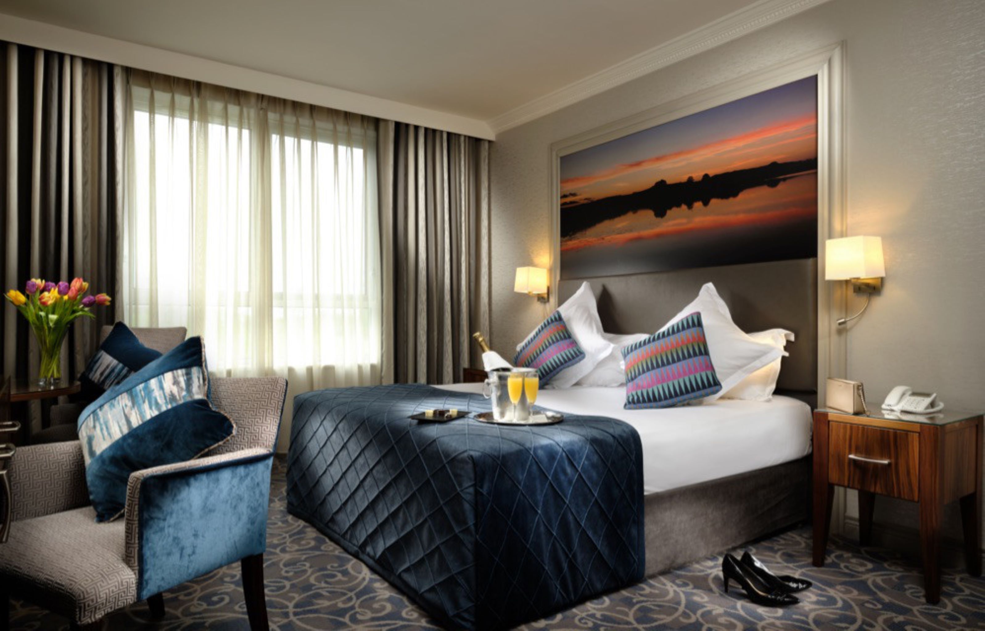 A hotel room with a bed, a chair, a nightstand, a lamp, and a painting on the wall.