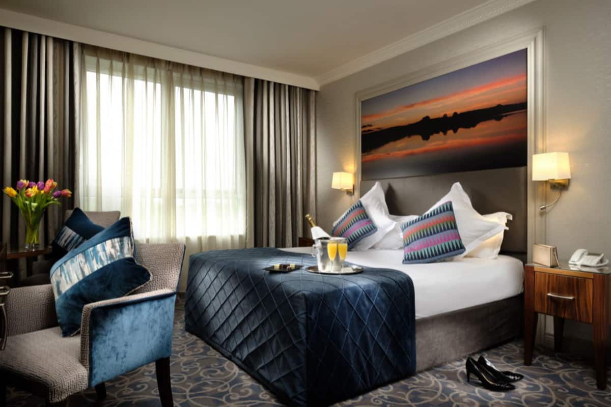 A hotel room with a bed, a chair, a nightstand, a lamp, and a painting on the wall.