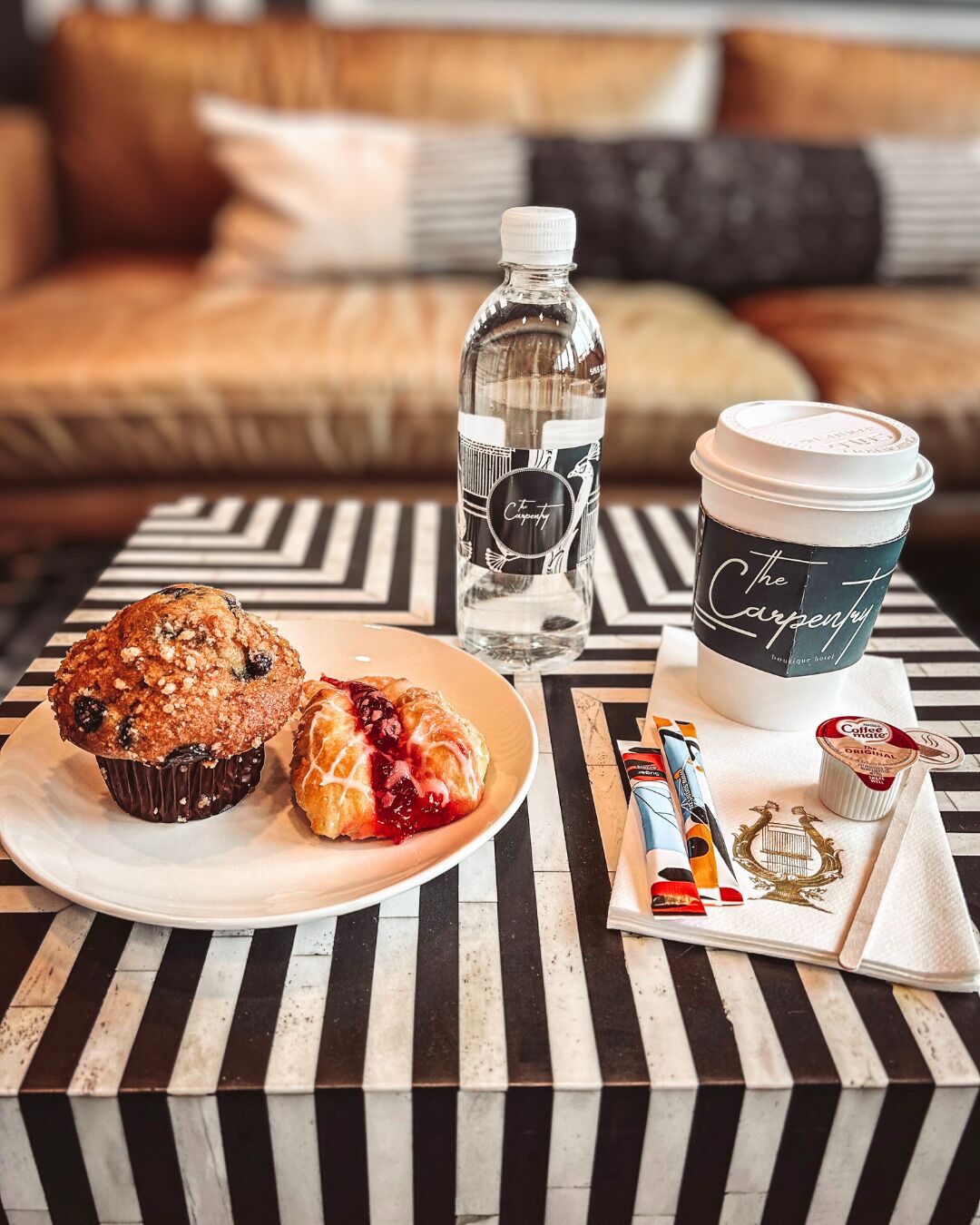 A table with a striped tablecloth, a plate with muffins, a bottle of water, a coffee cup, and a small tray with coffee creamer and a napkin.