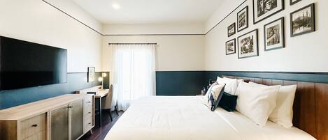 A hotel room with a large bed, a desk with a computer, a flat-screen TV, and a wall with framed pictures.
