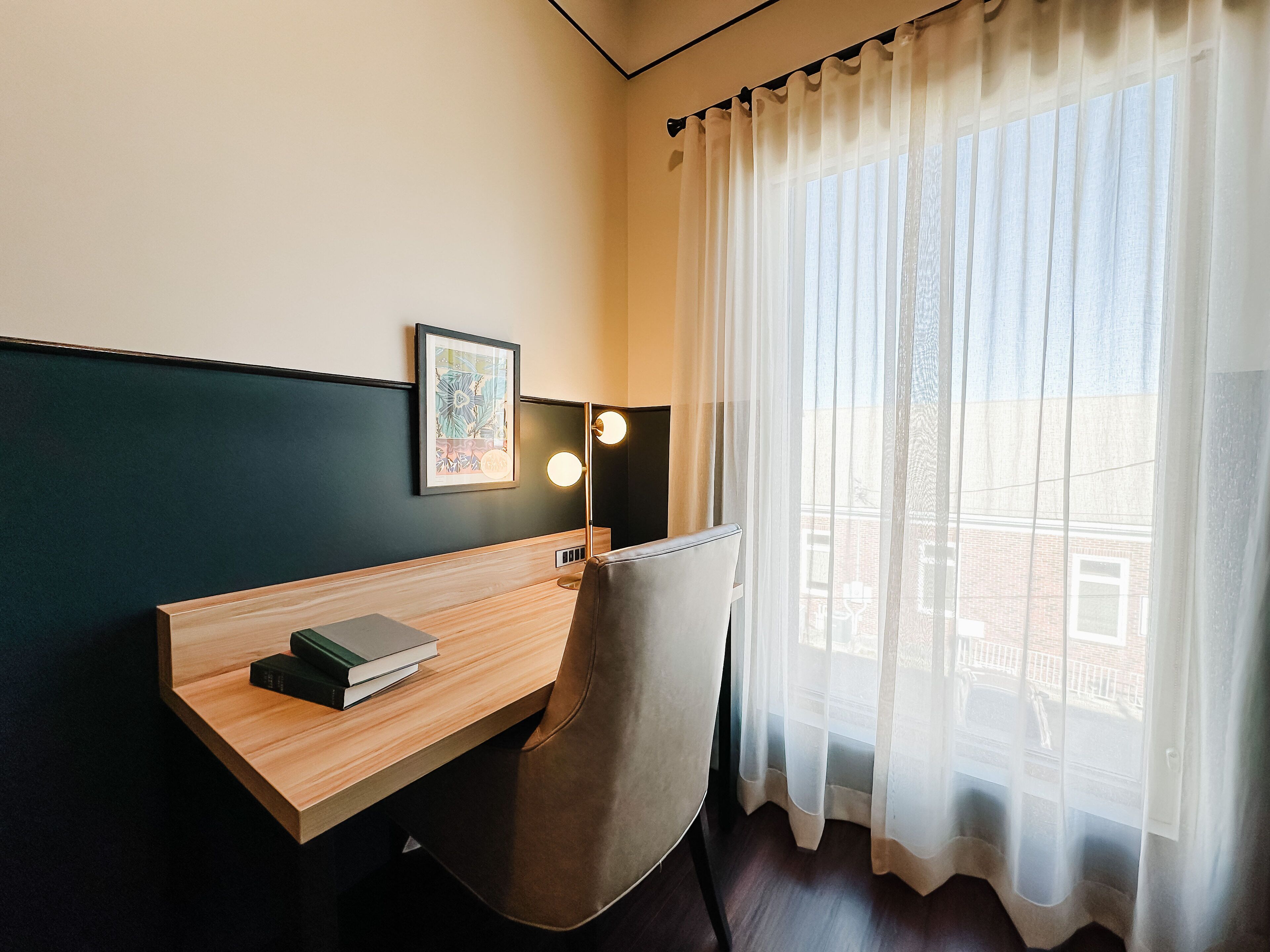 A hotel room with a wooden desk, a chair, a framed picture on the wall, and a window with sheer curtains.
