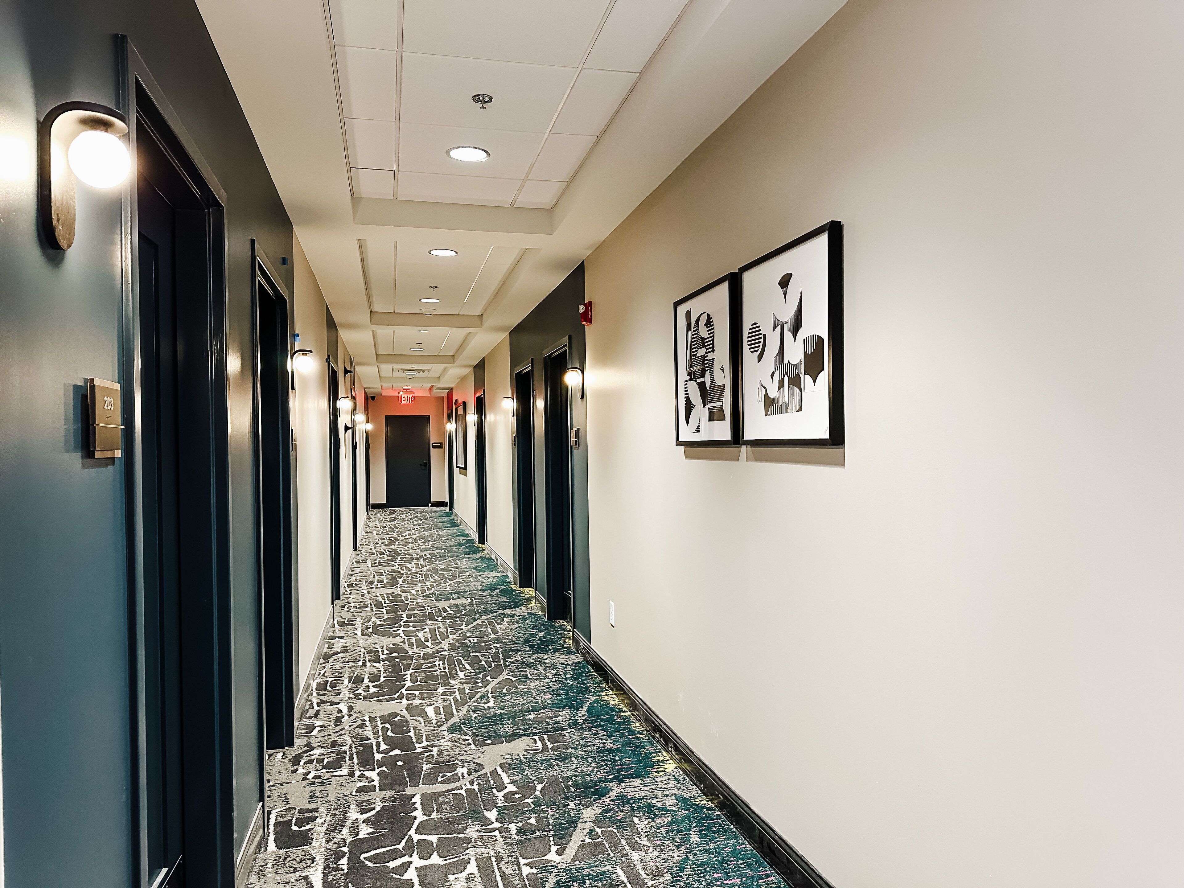 A hotel hallway with a patterned carpet, framed artwork on the walls, and doors on both sides.