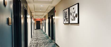 A hotel hallway with a patterned carpet, framed artwork on the walls, and doors on both sides.