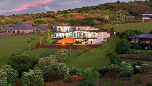 Uma casa grande e moderna, com vários níveis e um amplo jardim frontal, rodeada por vegetação exuberante e colinas.