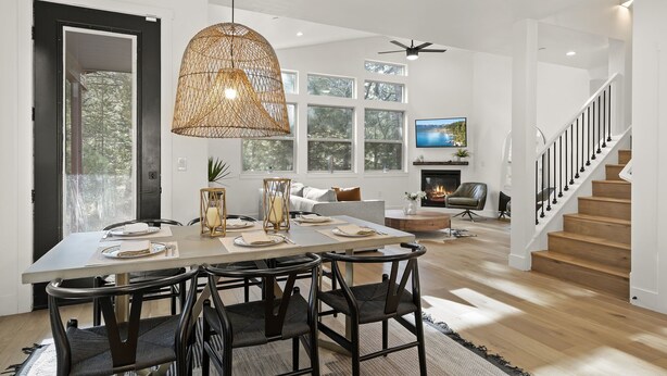 Un espace repas avec une grande table, des chaises et un luminaire suspendu. On y voit un escalier avec une rampe et, au fond, un téléviseur.