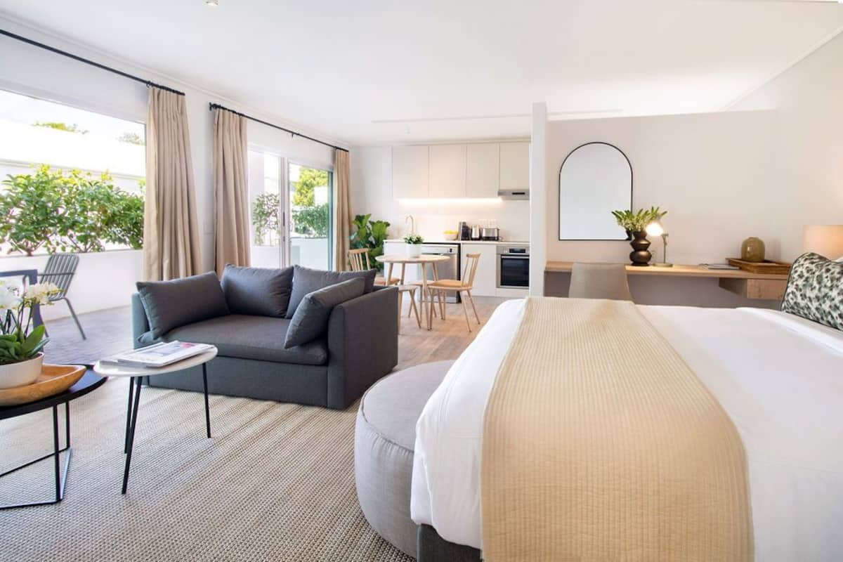 A modern hotel room with a sofa, a bed, a dining area, and a kitchen in the background.