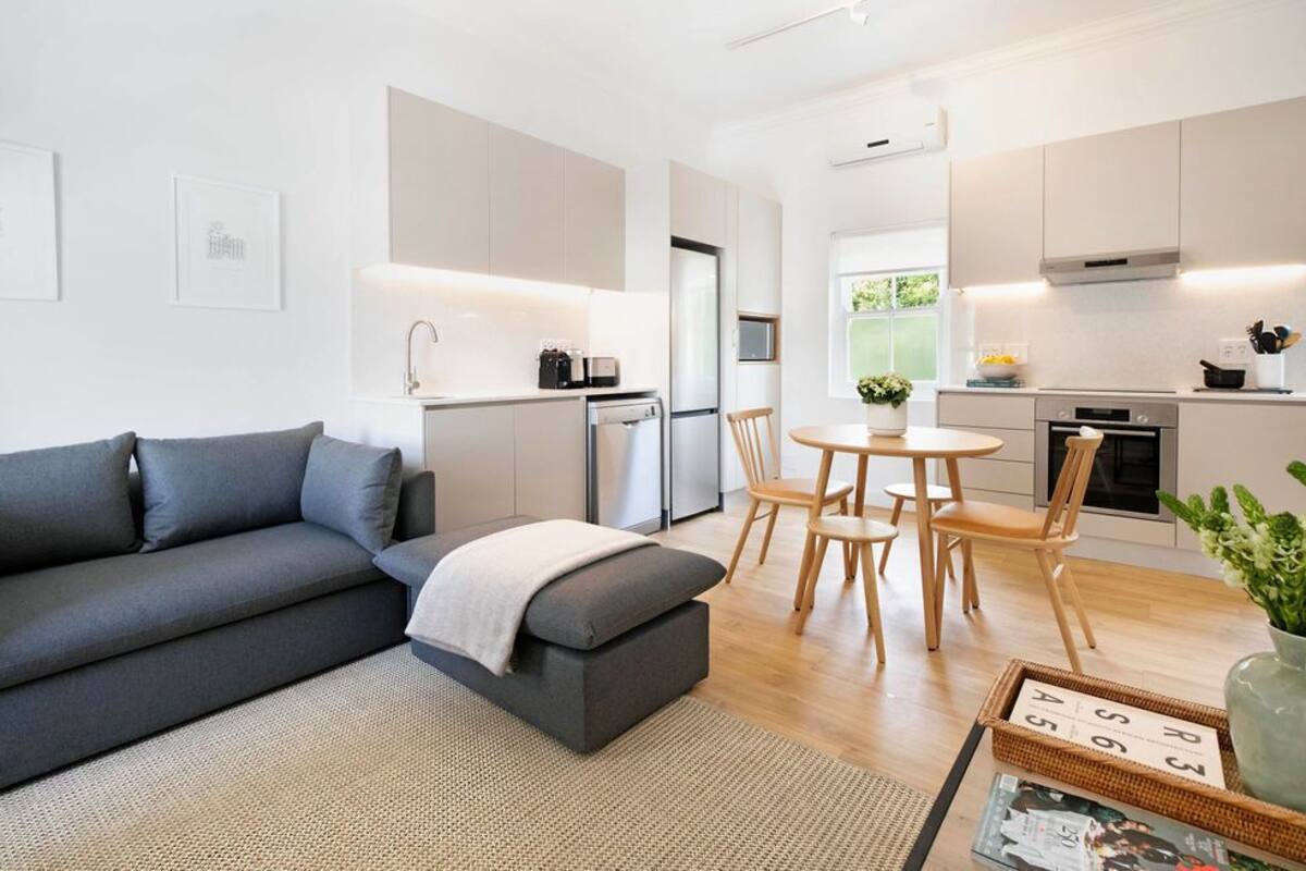 A modern living room with a grey sofa, a small table with chairs, and a kitchen area with a fridge, oven, and sink.