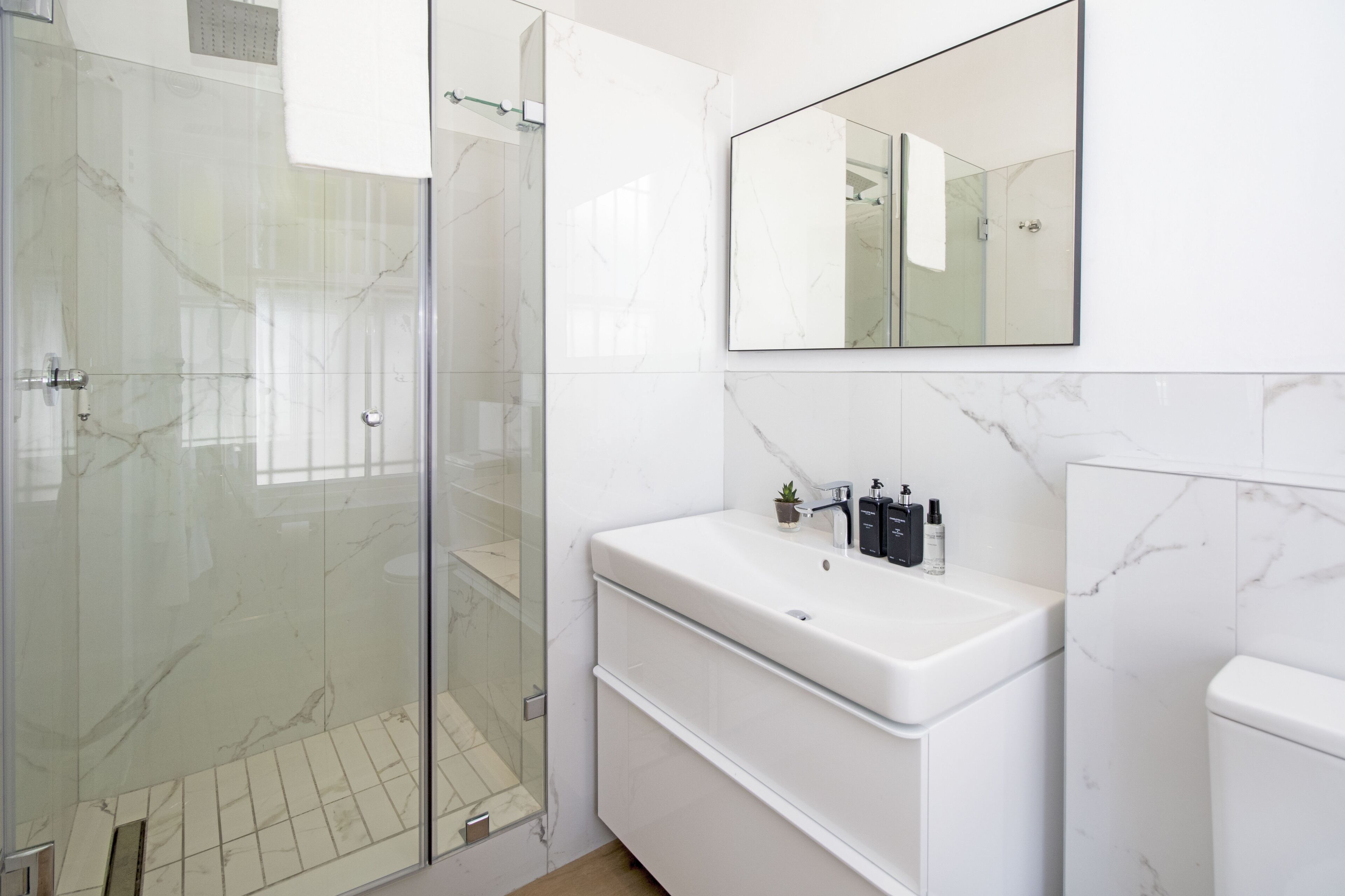 Een moderne badkamer met een glazen douchecabine, een witte wastafel met een marmeren blad en een grote spiegel boven de wastafel.