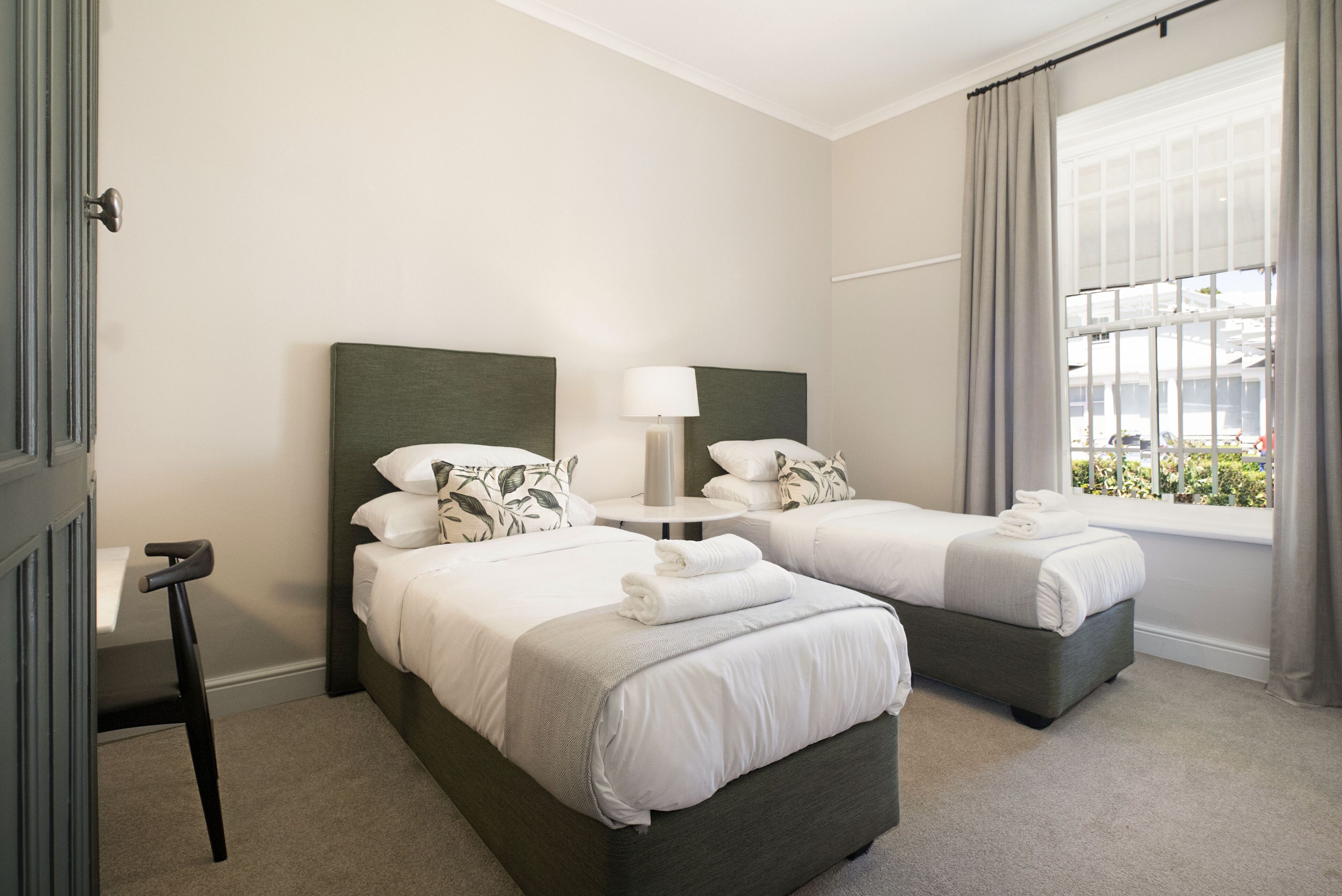 A hotel room with two beds, a bedside table with a lamp, a window with curtains, and a door.