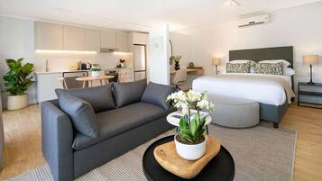 Un appartement moderne avec un canapé gris, une table basse en bois sur laquelle est posée une orchidée en pot, un lit avec une literie blanche, et une petite cuisine en arrière-plan.