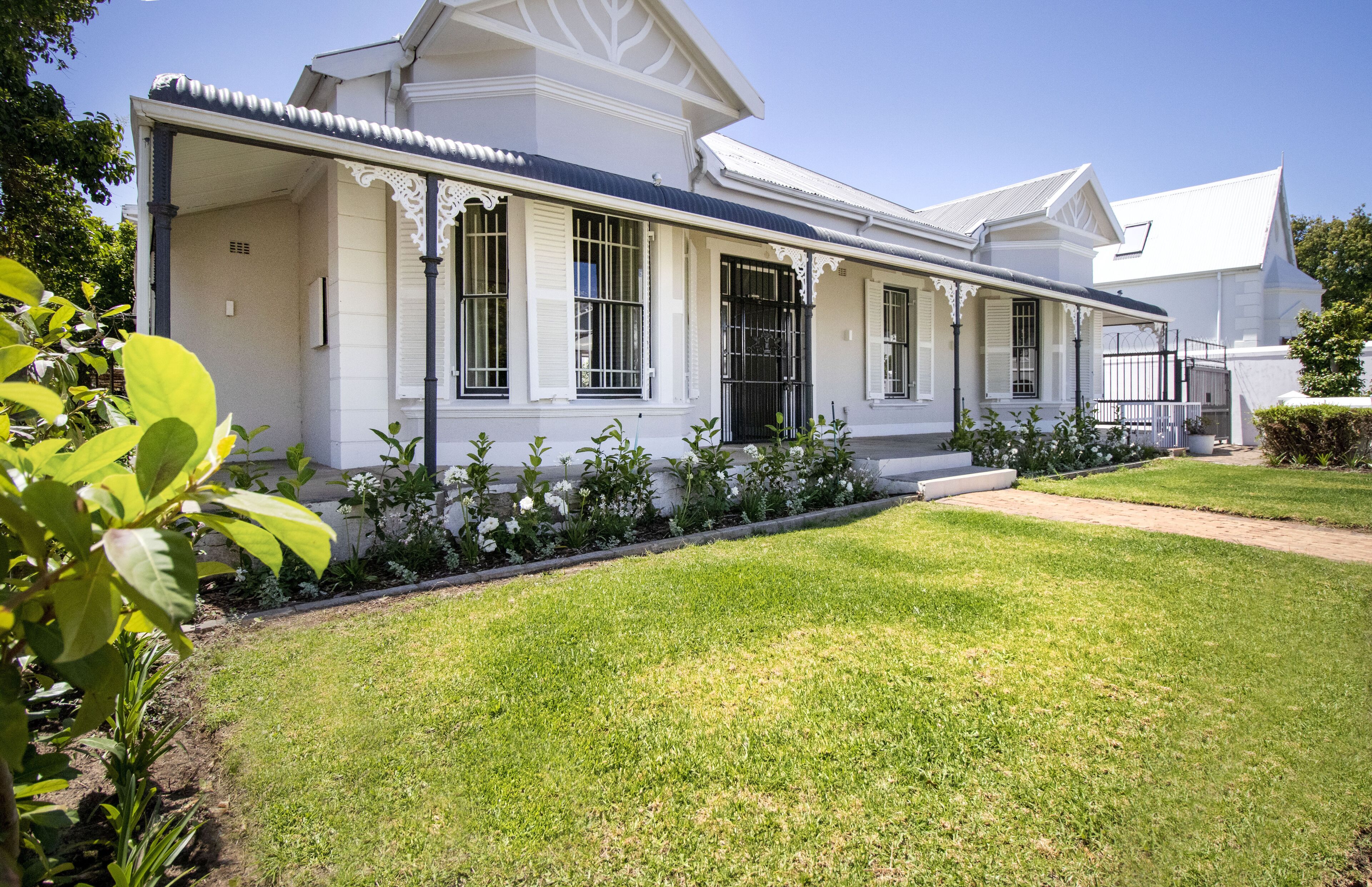 A white colonial-style house with a covered porch, a well-maintained lawn, and a garden with various plants.