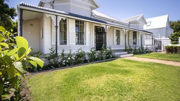 A white colonial-style house with a covered porch, a well-maintained lawn, and a garden with various plants.