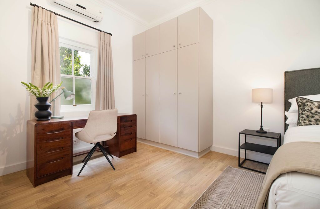 A bedroom with a bed, a desk with a chair, a wardrobe, a bedside table with a lamp, and a window with curtains.
