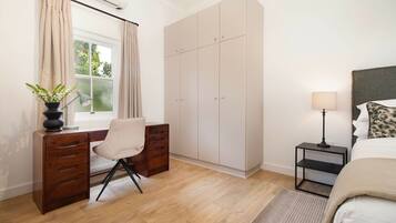 A bedroom with a bed, a desk with a chair, a wardrobe, a bedside table with a lamp, and a window with curtains.