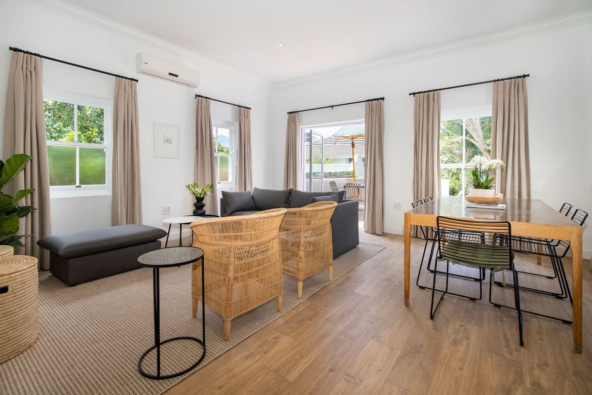 A modern living room with a sofa, two wicker chairs, a round table, and a dining table with chairs. There are large windows with curtains, a potted plant, and a small round table.