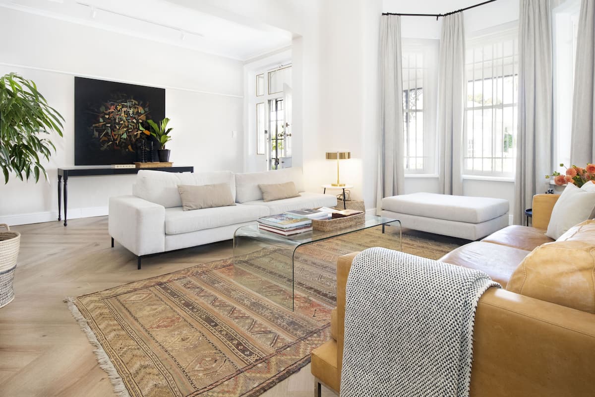 A modern living room with a white sofa, a glass coffee table, and a patterned rug. There is a large abstract painting on the wall and a potted plant on the left.